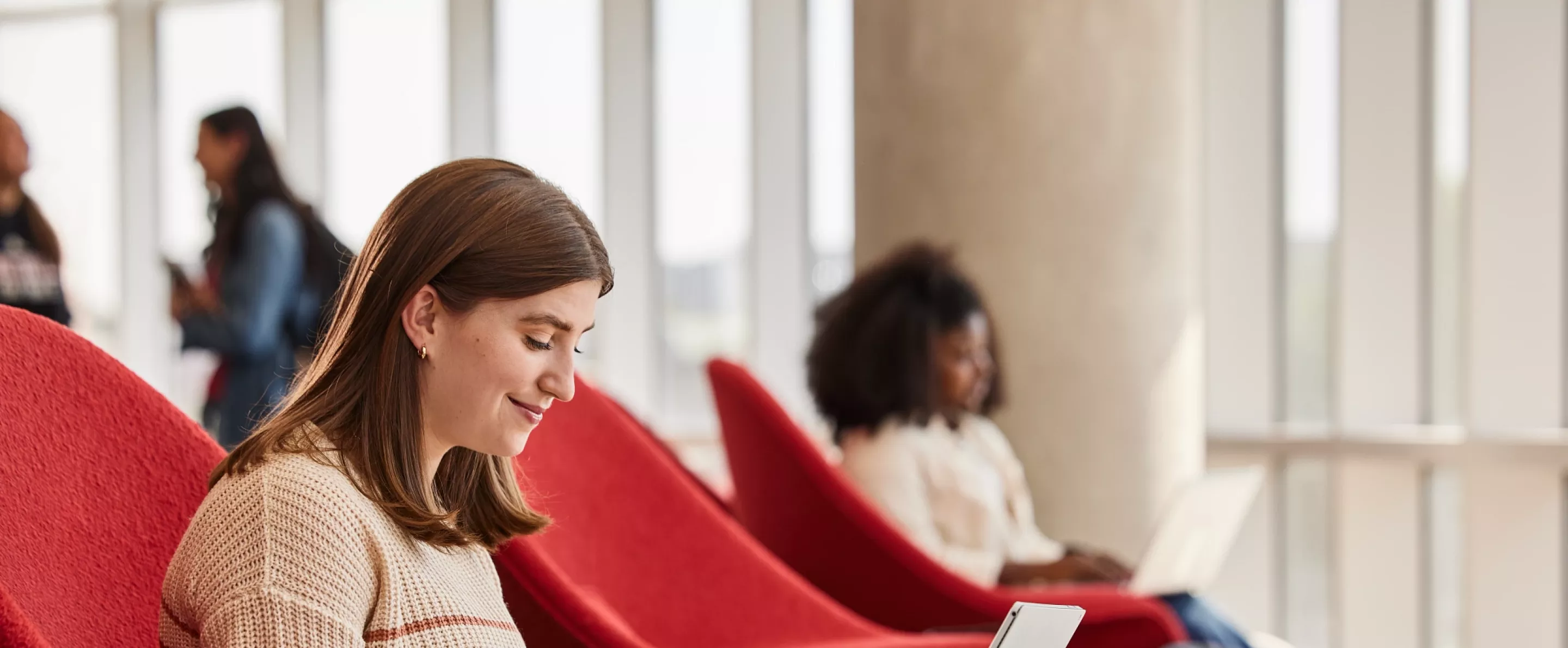 Lexi Raikes sitting on the second floor of the Belknap Academic Building, in a red chair, typing happily on her laptop