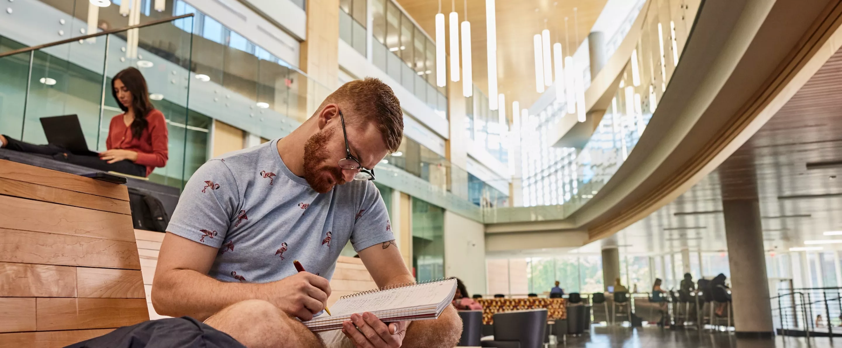 Student sitting with their legs crossed on the wooden platform benches in the Belknap Academic Building reading an academic textbook.