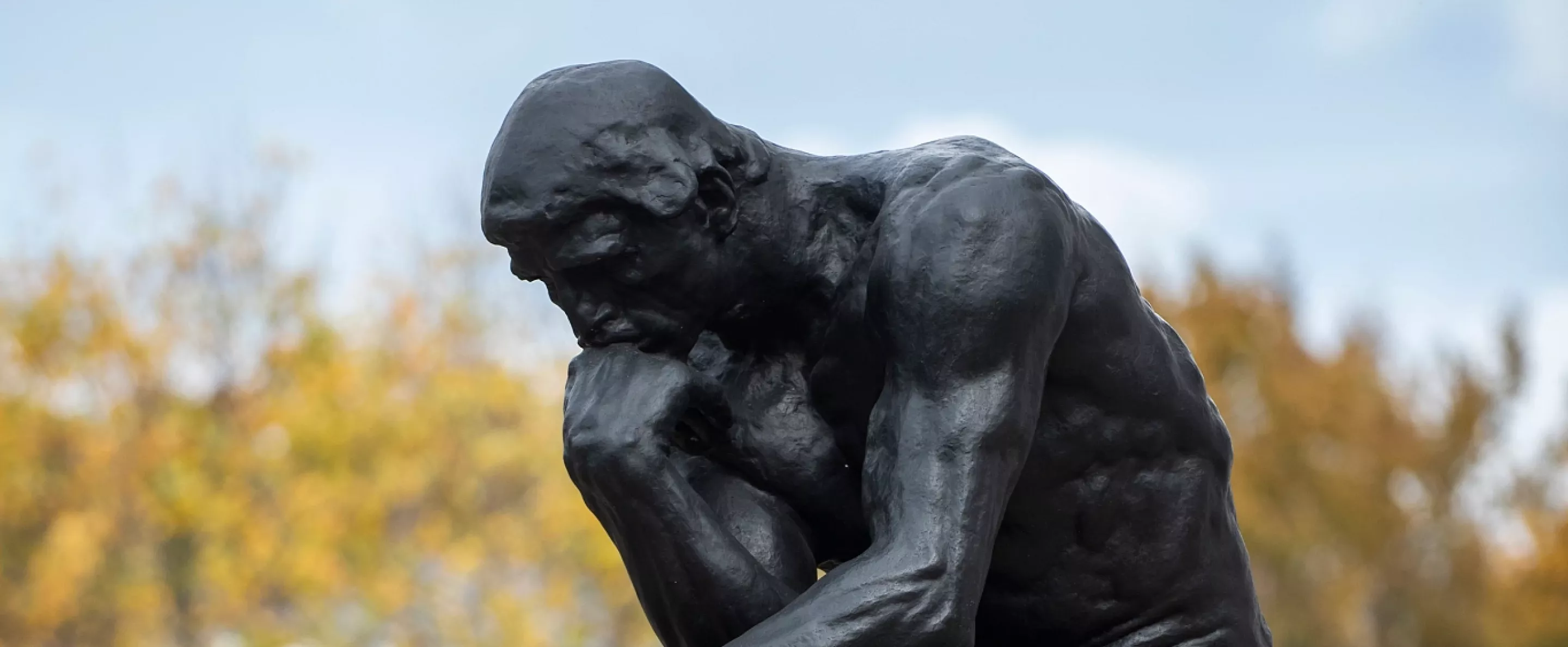 Side profile of The Thinker statue by Auguste Rodin outside of Grawemeyer Hall with fall foliage in the background