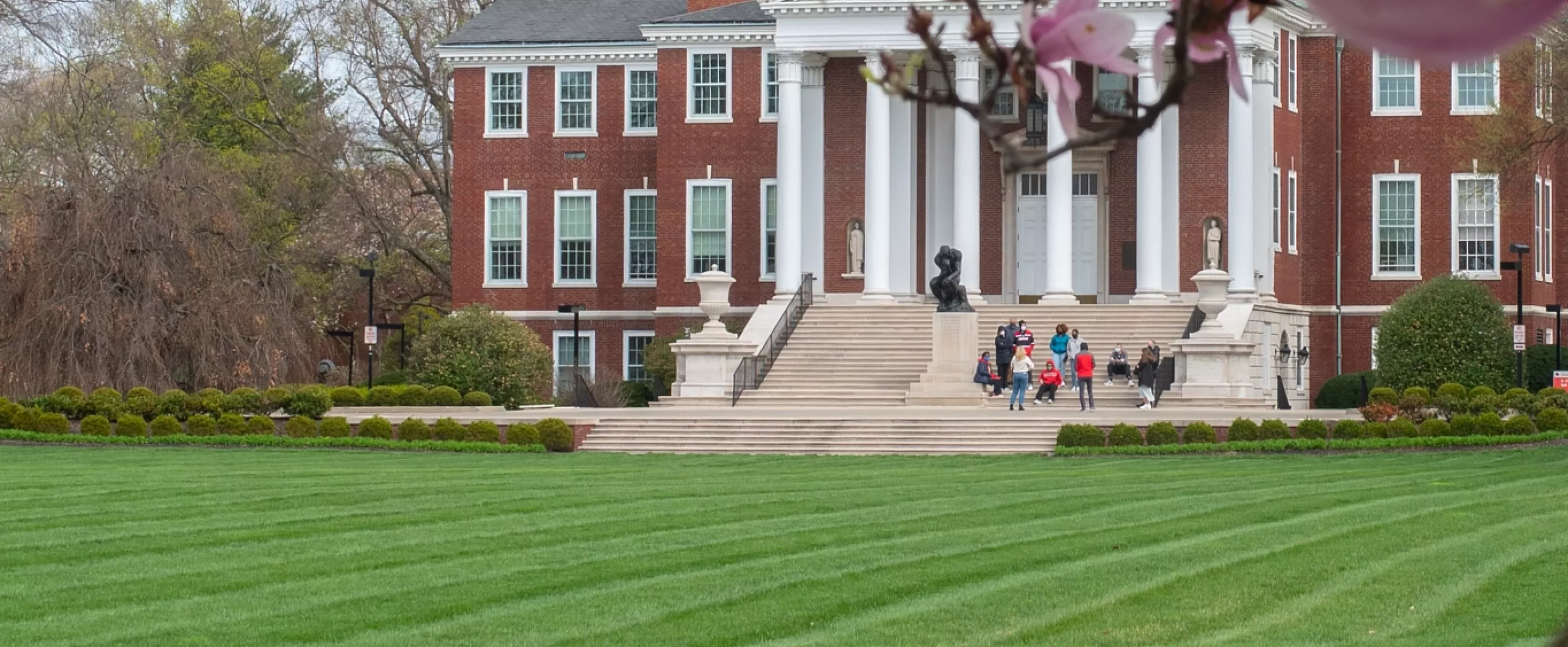 A group of people in the distance on the stairs of Grawmeyer Hall next to the Thinker statue, through spring blooms