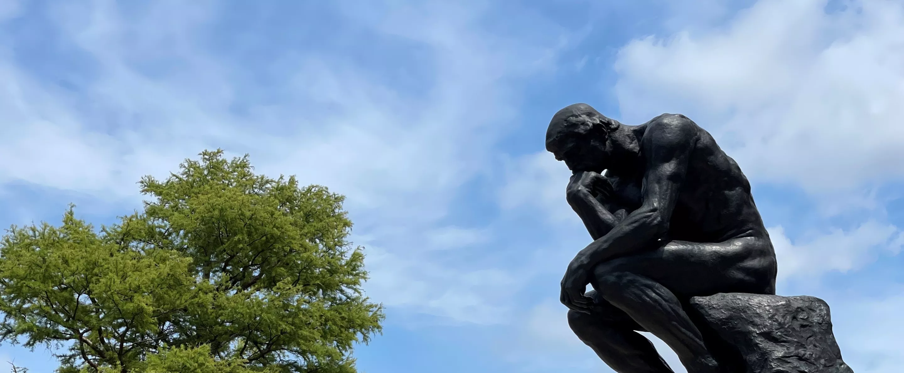 UofL's thinker statue from the side with the sky in the background. Trees on campus can be seen in the background as well as a lamp post with a UofL banner.