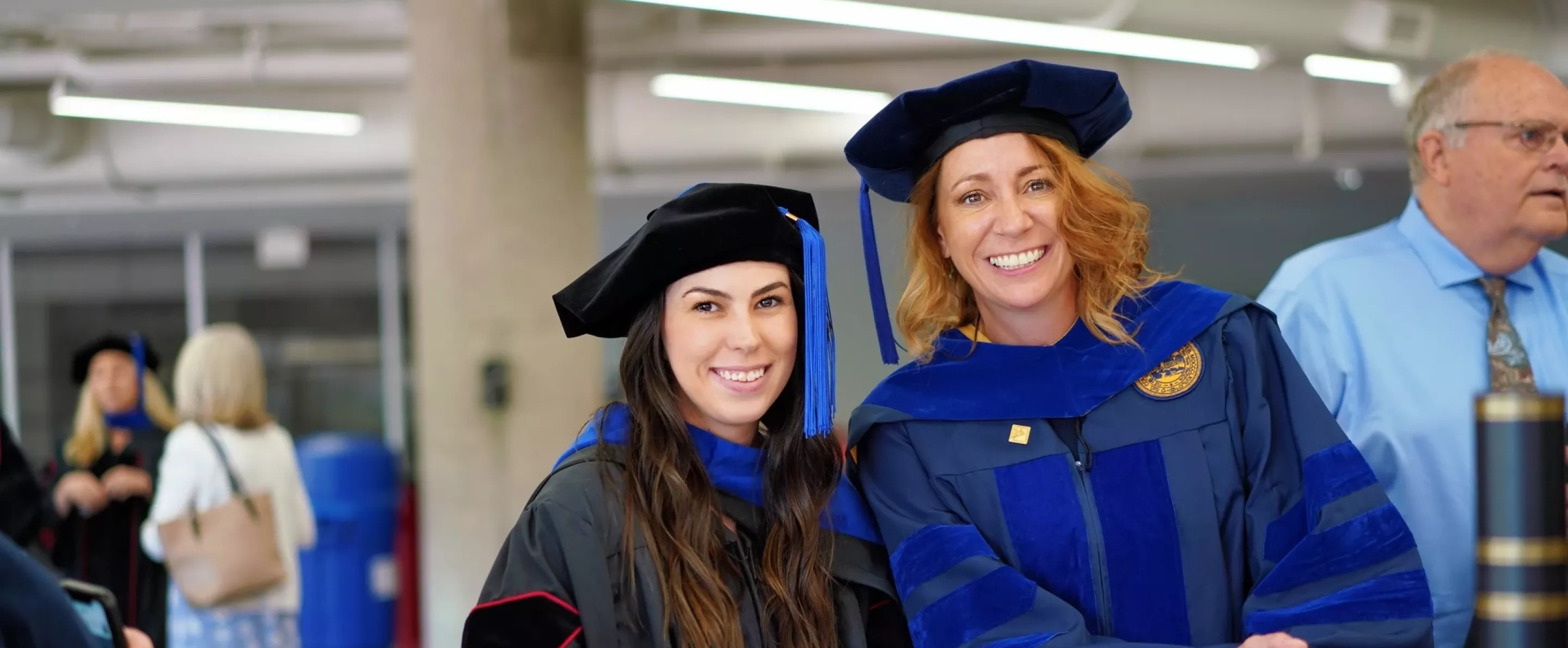 Two people standing near each other posing and smiling while in cap, gown and doctoral hood.