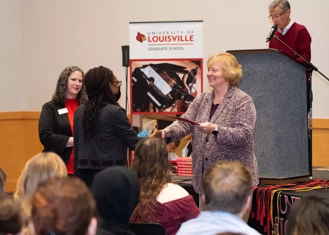 Person shaking hand and providing certificate to another person while in front of a seated crowd.